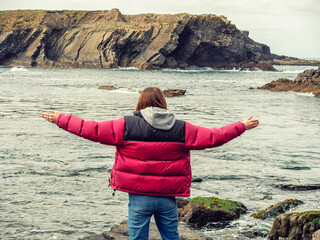 Fototapete - A woman overlooking the ocean and rough stone coast. She is wearing a red jacket and blue jeans. The scene is peaceful and serene, with the ocean as a backdrop. Bridges of Ross area, Ireland travel