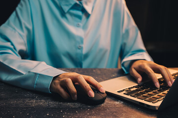 Poster - businessman working. Professional person in blue shirt using laptop with computer mouse on wooden table in low light setting