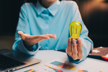 Poster - Idea concept. Person Holding Yellow Bulb While Explaining Ideas with Charts and Laptop on Table in Bright Office Environment