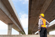 © tong2530 - An engineer, wearing a protective helmet and safety vest, inspects the structural integrity of a concrete overpass against a clear, bright sky.