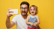 © Vasiliy - Excited father and curly child taking selfie wearing funny glasses on yellow background. Joyful family photography moment. Father's Day camera and entertainment service promotions