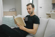 © APchanel - Man seated on sofa reading book and drink a cup of coffee or tea mood is peaceful and introspective, capturing a moment of personal retreat in a well-designed, comfortable environment.