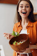 © SHOTPRIME STUDIO - Happy young woman enjoying a healthy salad in a wooden bowl, wearing a bright orange shirt, set in a modern kitchen with a warm color palette