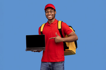 Poster - Order Here. Portrait of smiling black delivery man holding laptop and pointing at empty screen for mockup, wearing red uniform and yellow thermo backpack, standing on blue studio background, copyspace