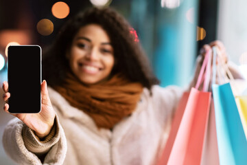 Poster - Online Shopping Concept. Young smiling african american woman showing smartphone screen with black blank empty screen with mockup for advertisement, holding bags, selective focus, blurred background
