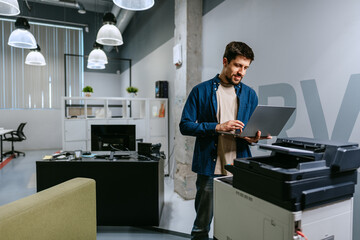 Poster - Young man using laptop while standing next to printer in modern office setting during daytime
