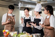 © bongkarn - Asian female chef holding a tablet while her students looking at seasonal jars over kitchen counter.