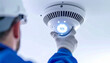 © Tossakan Photos - Worker installing a modern smoke detector with bright LED lights on a white ceiling for enhanced safety.