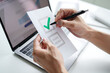 © mojebor - Close-up of a person's hands filling out a checklist on paper with a green check mark over the first option while using a laptop for reference.