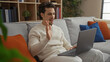 © Krakenimages.com - Young hispanic man sitting on a sofa in a cozy living room using a laptop for a virtual meeting, with bookshelves and plants in the background, appearing engaged and smiling.