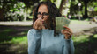 © Krakenimages.com - Middle-aged woman in park holding argentine pesos, wearing glasses and sweater, expressing anger with trees and sunlight in background.