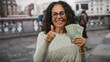 © Krakenimages.com - Middle-aged woman in uae holds dirham banknotes and gives thumbs-up on a bustling city street.