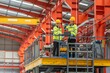 © BESTIMAGE - Industrial workers wea safety helmets and high visibility jackets inspecting equipment on an elevated platform inside a large factory or warehouse with orange steel beams and overhead cranes