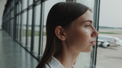 Poster - Professional businesswoman standing near airport window, gazing thoughtfully at airplanes while contemplating upcoming business journey and personal aspirations