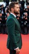 © Mid - Elegant man in dark green suit posing on red carpet looking away from the camera during a daytime event. Professional attire with blurred background and many people.