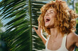 © SHOTPRIME STUDIO - joyful woman with curly hair laughing outdoors near tropical palm leaves in bright sunlight, expressing happiness and natural beauty in summer atmosphere.