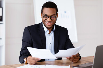 Cheerful young black manager in glasses reading marketing report and smiling, sitting at his workplace in front of laptop in office, african american businessman doing paperwork, copy space