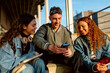 © Alberto - Three cheerful students are sitting on steps outside, sharing content on a smartphone and enjoying their time together