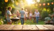 © Vadym - Empty wooden table in front of blurred BBQ party scene. Group of people enjoy summer picnic outdoors, grill on green background. Wooden deck table, natural green garden backdrop. BBQ concept.