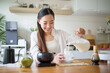 © matinee - Smiling woman pouring milk into a glass to prepare iced matcha latte in a kitchen.