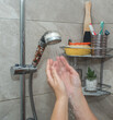 © fototema - Close-up of hands under a stream of water. The shower head has a transparent handle with filter granules. A shelf with bath accessories is visible in the background.