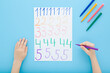 © fotoduets - Little child boy hand holding color pen and writing colorful different numbers on white paper on blue table background. Pastel color. Doing school exercise. Closeup. Point of view shot. Top down view.