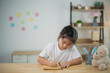 © WMSTUDIO - Young girl with black hair writing on paper at a wooden table in a cozy indoor environment surrounded by toys and colorful post-it notes on the wall