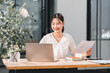 © kenchiro168 - Young Asian woman working at desk with laptop, holding business report, smiling in modern office, natural light, professional and productive atmosphere