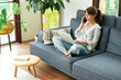 © UnitedPhotoStudio - A woman comfortably works from home on a couch, using her laptop and talking on the phone, surrounded by plants and natural light.
