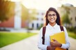 © EdNurg - Smiling Indian student holding books and a backpack, enjoying a sunny day on the university campus, embodying the spirit of education and youth