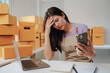© Tj - Young asian woman is having problems with her online business and is looking at her smartphone while holding her head with her hand, surrounded by cardboard boxes, a laptop and a notebook