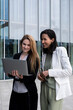 © yaqui_villegas - Two women in their 30s from different ethnicities collaborating outdoors on a laptop in a modern corporate setting