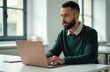 © Pete - Bearded man works on laptop at desk in bright office. Business pro looks at computer screen. Male in green sweater. Remote work, online communication, modern workplace, corporate environment.