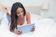 © wavebreak3 - African American woman lying on bed in bedroom using tablet with white bedding and table lamp
