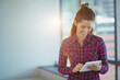 © WavebreakMediaMicro - Woman in plaid shirt standing near large windows tapping tablet in office sun flare, copy space