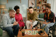 © Seventyfour - Diverse group of young adults including Black woman and Caucasian man with disability throwing dice sitting together playing board game at birthday celebration, drinks and game pieces on table
