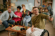 © Seventyfour - Diverse group of young adults including Black woman and Caucasian man with disability in wheelchair celebrating birthday, playing board game, smiling and taking selfie using selfie stick at home