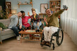 © Seventyfour - Group of young adults including Black woman, Caucasian man, Caucasian woman, and man with disability in wheelchair taking selfie using selfie stick and celebrating birthday together in living room