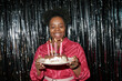 © Seventyfour - Portrait of adult Black woman smiling while holding birthday cake with lit candles in front of metallic backdrop, celebrating birthday and looking at cake
