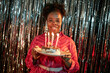 © Seventyfour - Portrait of young adult Black woman smiling and holding birthday cake with lit candles in front of metallic backdrop celebrating birthday in festive setting