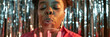 © Seventyfour - Website header shot of Black woman blowing out candles on birthday cake celebrating holiday while standing against metallic tinsel background