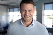 © wavebreak3 - Man in his 30s wearing light-blue sweater smiling at camera in office with desks, large windows