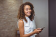 © WavebreakMediaMicro - African American woman in white blouse standing while smiling, holding smartphone in office hallway