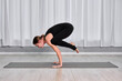 © TRAVELARIUM - Young, caucasian woman in black outfit performs crow pose on mat in minimalist studio with white curtains. Scene exudes focus and calm, highlighting balance and strength