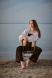 © TRAVELARIUM - Young woman in casual attire sits on chair at serene beach, embodying calmness and introspection. Soft evening light and water create peaceful, reflective mood