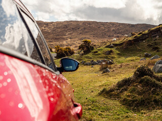 Wall Mural - A red family car is parked on a road by a field. The car is parked on a dirt road with a few trees in the background. The sky is cloudy. Travel and rural area tourism