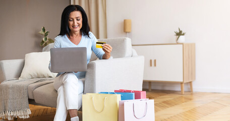 Poster - Buying Gifts And Presents Concept. Happy Mature Lady Using Laptop, Holding Credit Card, Ordering Goods In Internet Store, Sitting On The Couch At Home, With Shopping Bags On The Floor, Copy Space