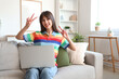 © Pixel-Shot - Beautiful young woman in rainbow t-shirt with modern laptop showing victory gesture and sitting on sofa in living room. LGBT concept
