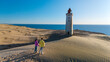 © Fokke Baarssen - A couple stands hand in hand on the sandy dunes near Rubjerg Knude lighthouse, overlooking the breathtaking coastline of Denmark during a golden sunset.