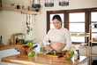 © bongkarn - Pregnant asian woman holding knife cutting cucumber on chopping board preparing her food in kitchen.